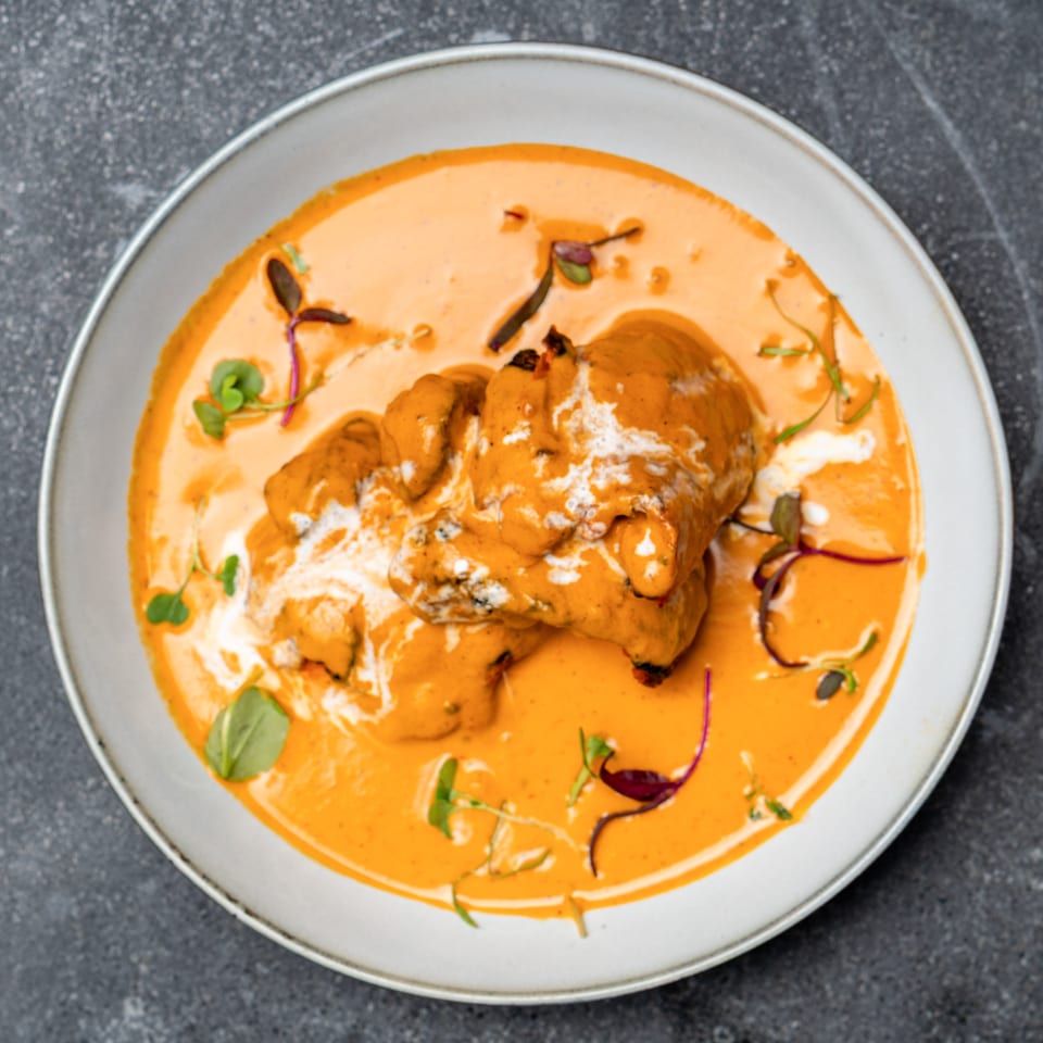 Overhead view of grilled chicken pieces in creamy orange sauce, garnished with microgreens and drizzled cream on a white plate.