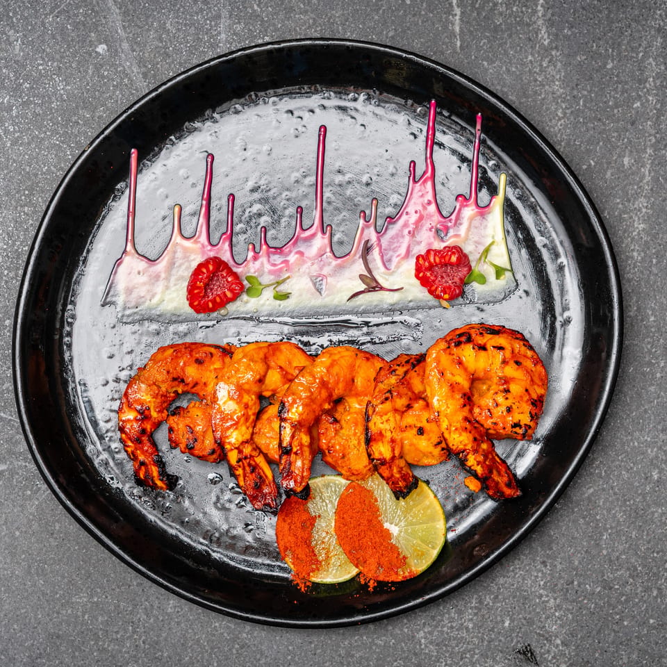 Black plate of grilled orange prawns with charred marks, lime, chili powder, microgreens, raspberries, and pink sauce.