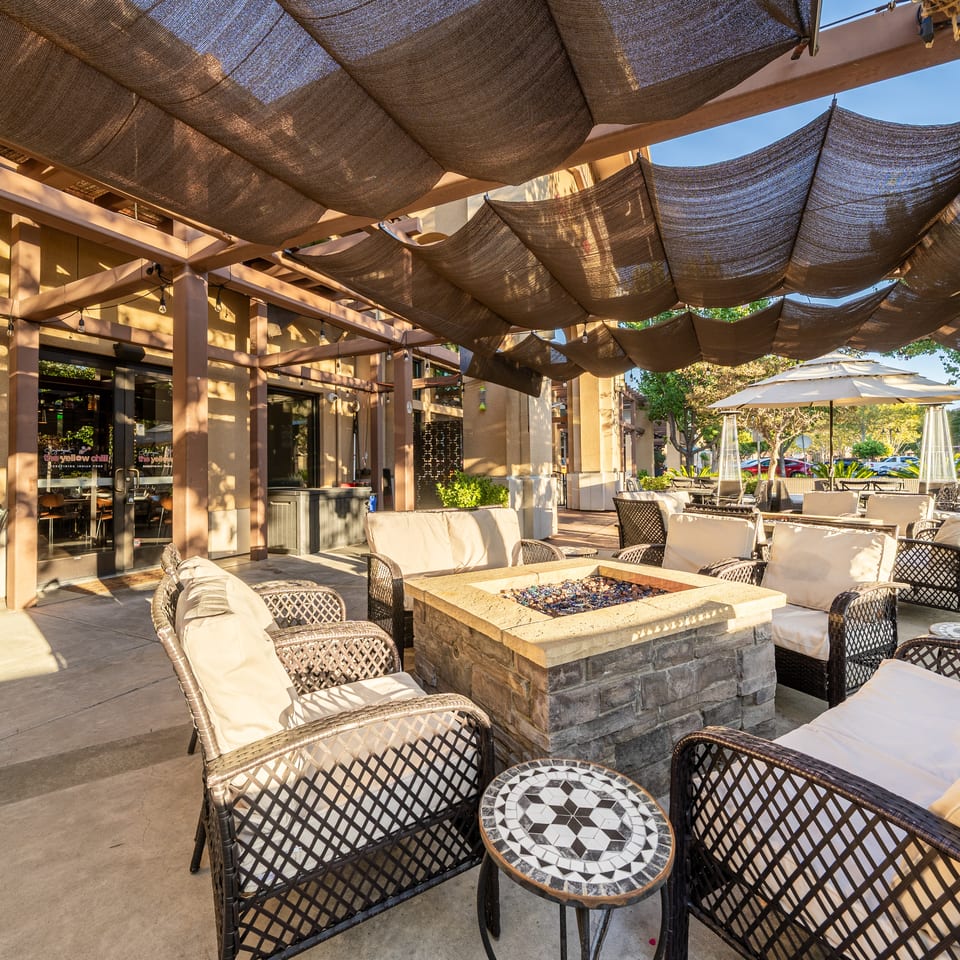 Outdoor restaurant patio with cushioned wicker seating, mosaic tables, fire pit, umbrellas, and shade sails.