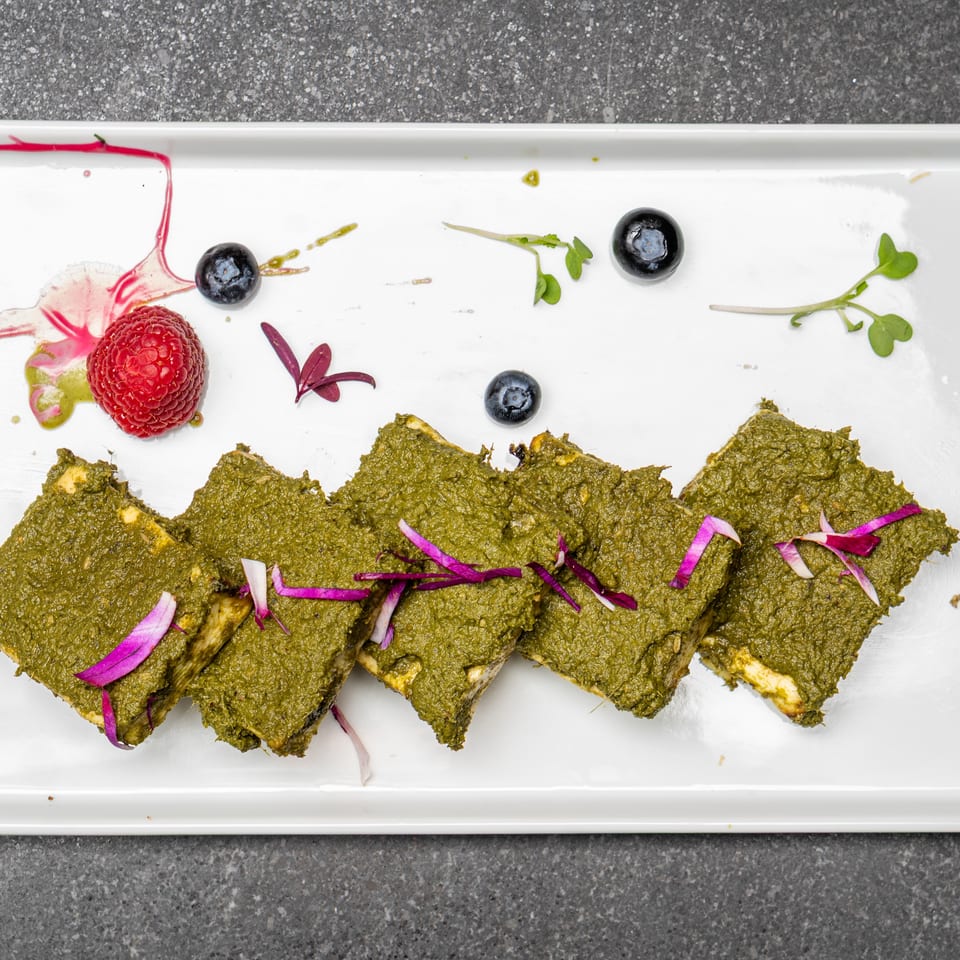 Green herb-coated paneer slices arranged on a white platter with blueberries, raspberry, microgreens, and colorful sauces.
