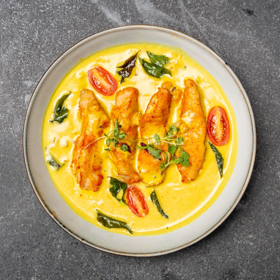 Golden curry with seared fish, cherry tomatoes, curry leaves, and microgreens served on a ceramic plate.