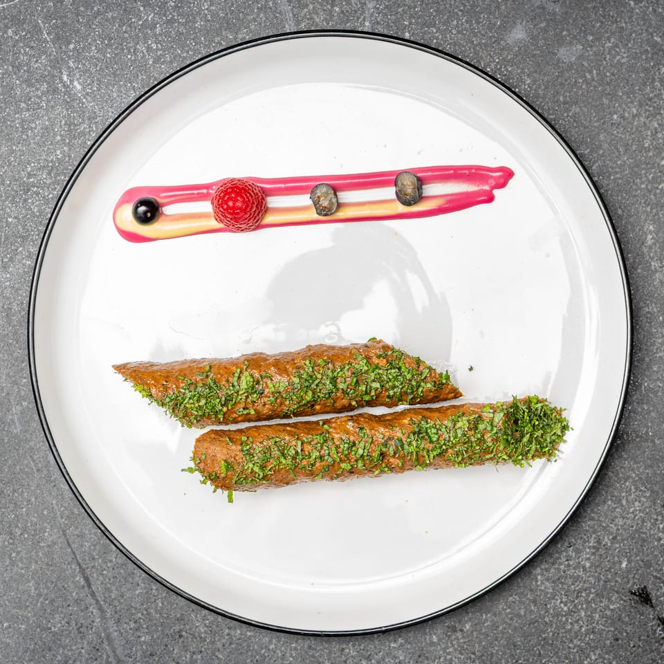 Two herb-coated grilled kebabs arranged on a white plate with raspberry, blueberries, and colorful sauce streaks.