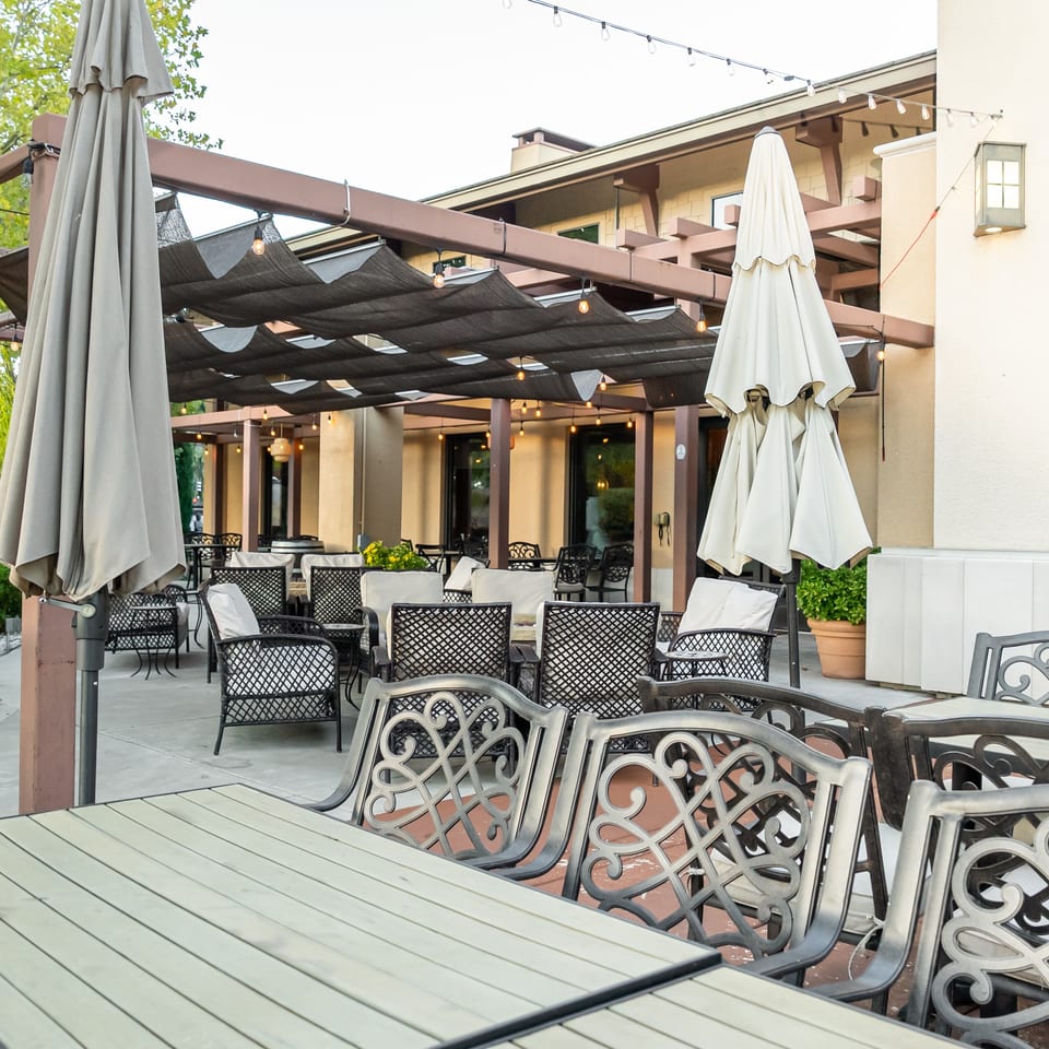 Outdoor restaurant patio with ornate chairs, tables, closed umbrellas, pergola shade, and string lights.