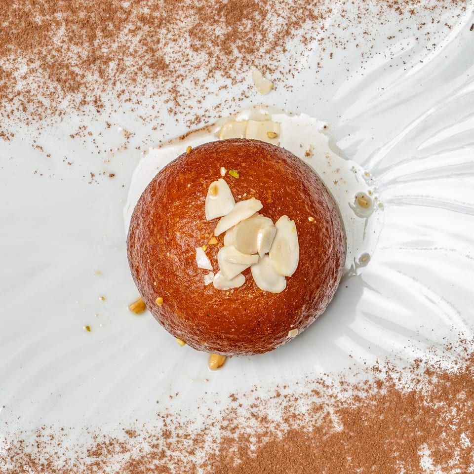 Round glazed pastry topped with sliced almonds, pistachios, and cream on a white plate dusted with cocoa powder.