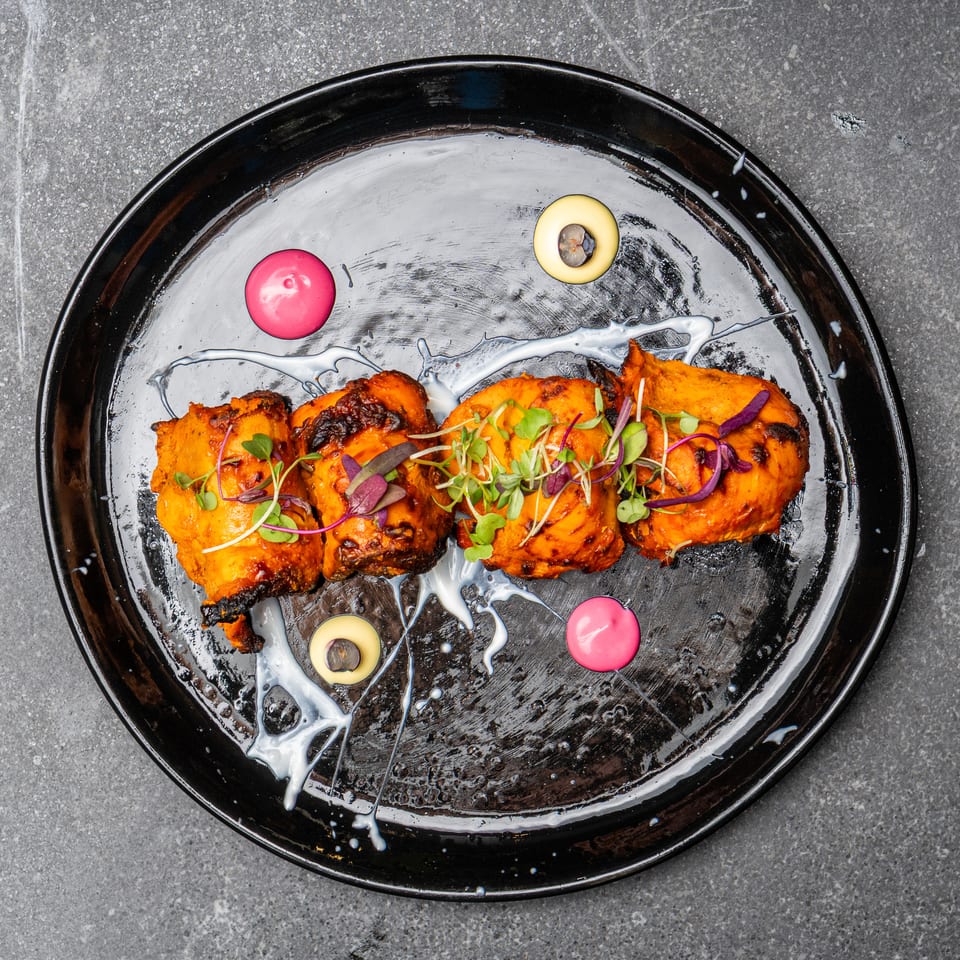 Grilled orange-marinated chicken pieces topped with microgreens on a black plate, drizzled with creamy sauce and colorful purées.