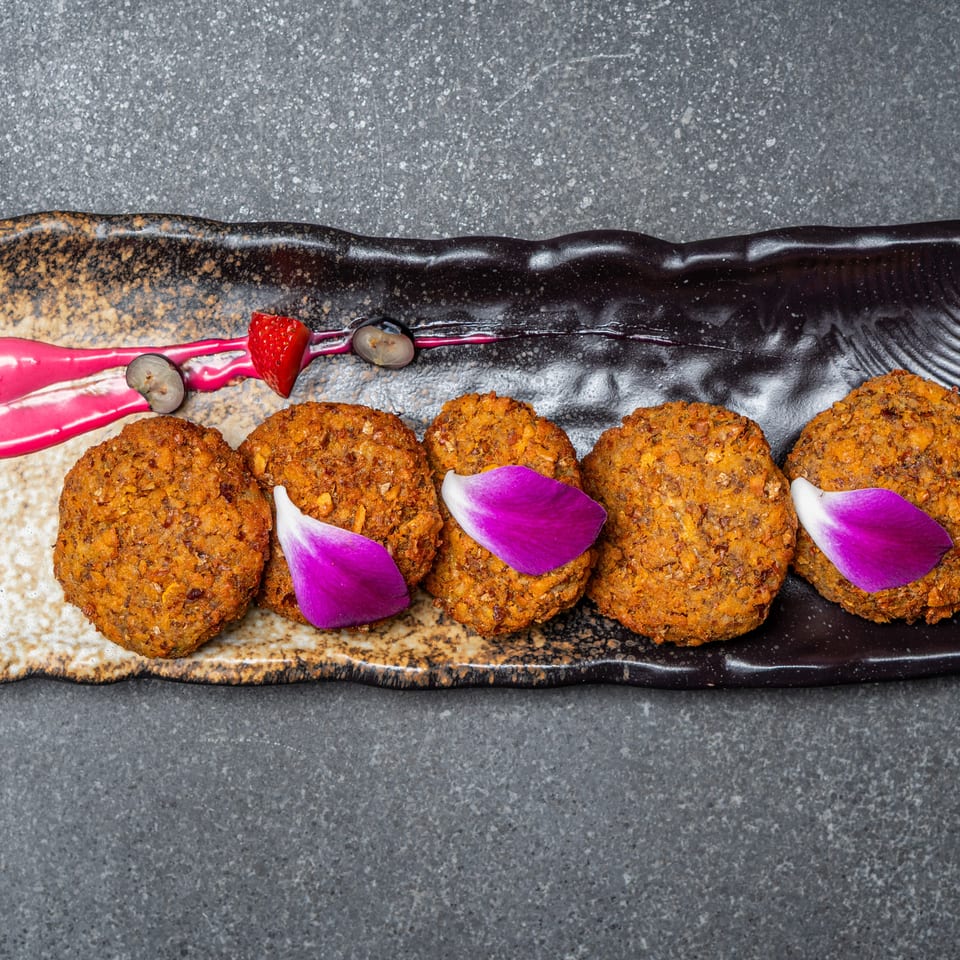 Five golden-brown croquettes arranged on a black serving plate with pink sauce, berries, and purple flower petals.